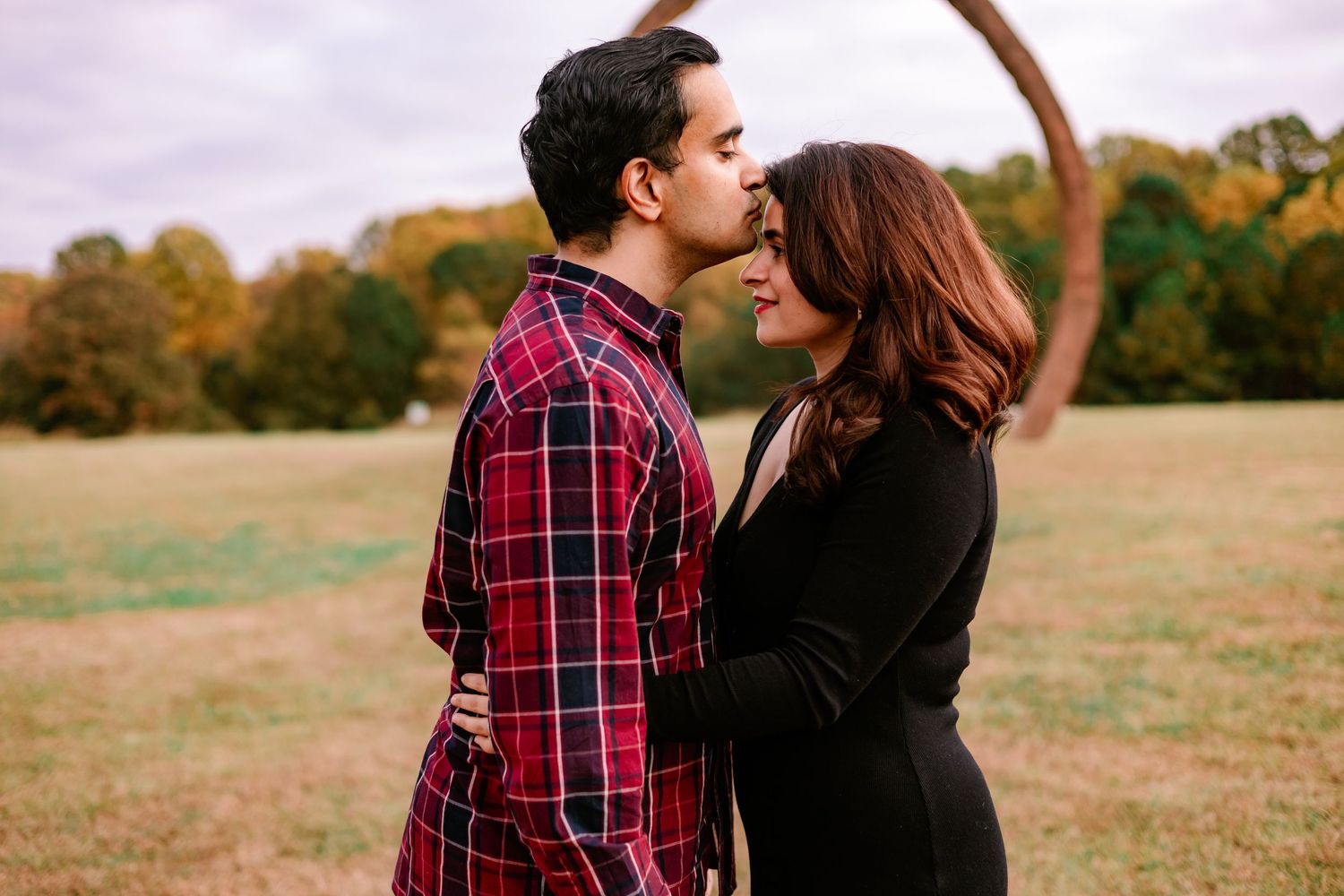 Couple sharing a tender kiss in an open field with autumn trees in background.