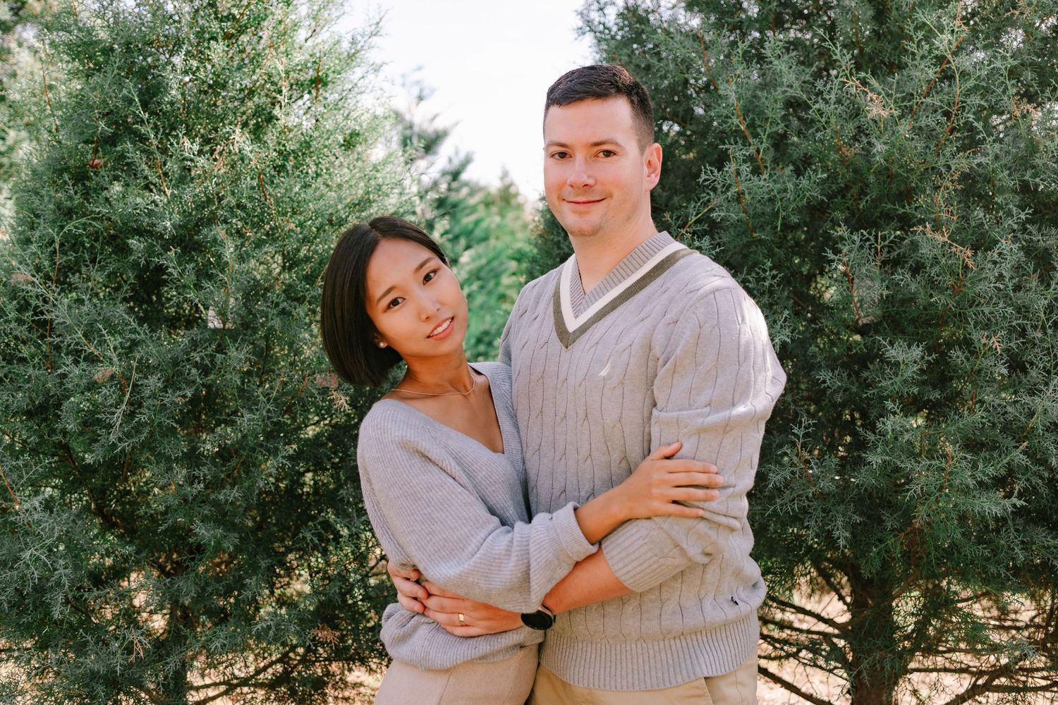 Smiling couple embracing in front of lush evergreen trees in soft natural light.