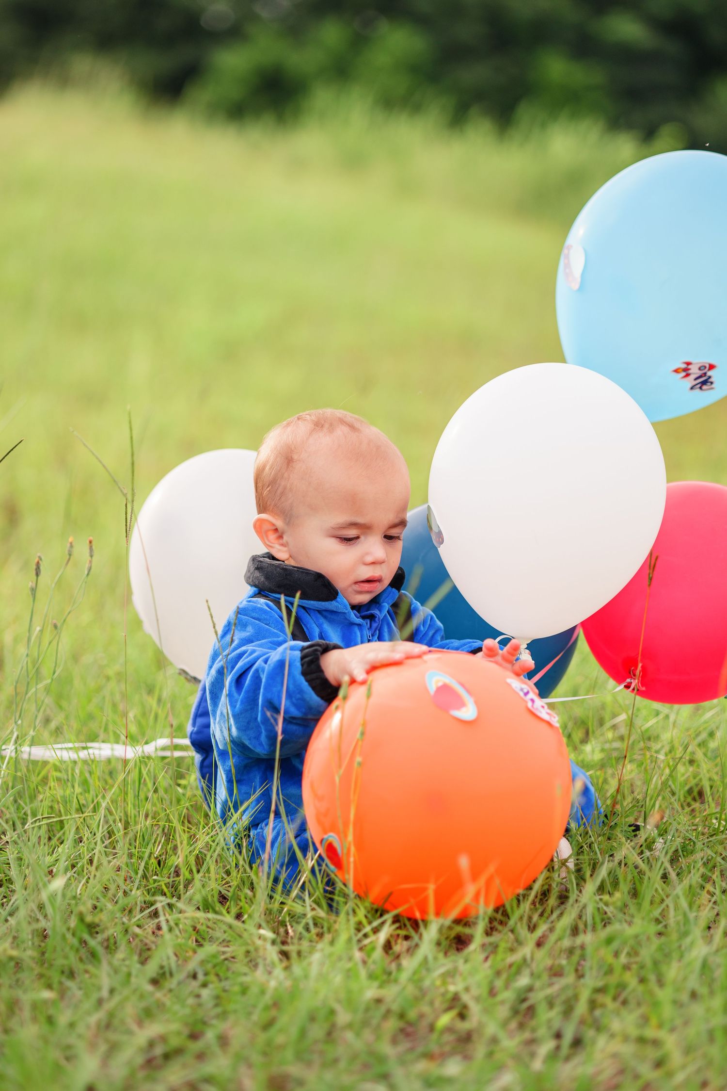 A small child in a blue spacesuit plays with colorful balloons in tall grass.