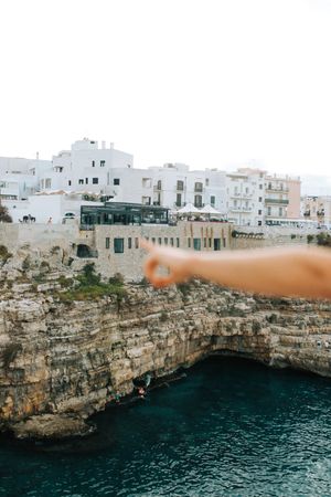 A hand points toward white buildings perched on rocky cliffs above turquoise Mediterranean waters in Italy.