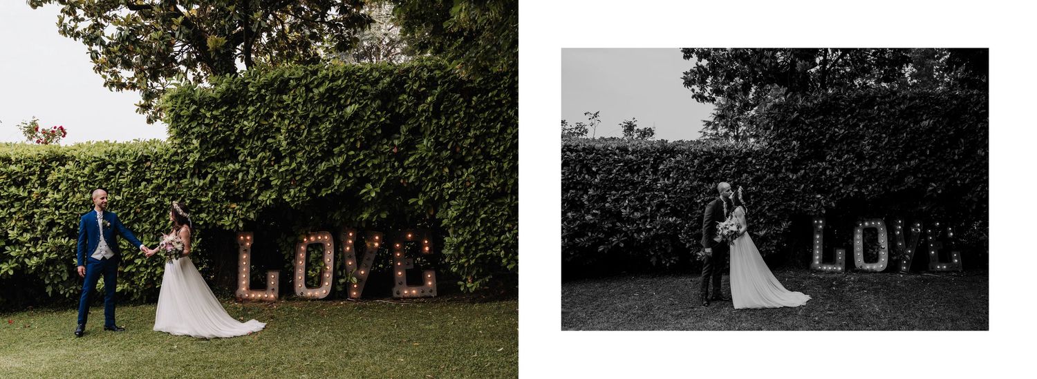 Bride and groom posing in front of large LOVE letter lights in a lush garden wedding setting.