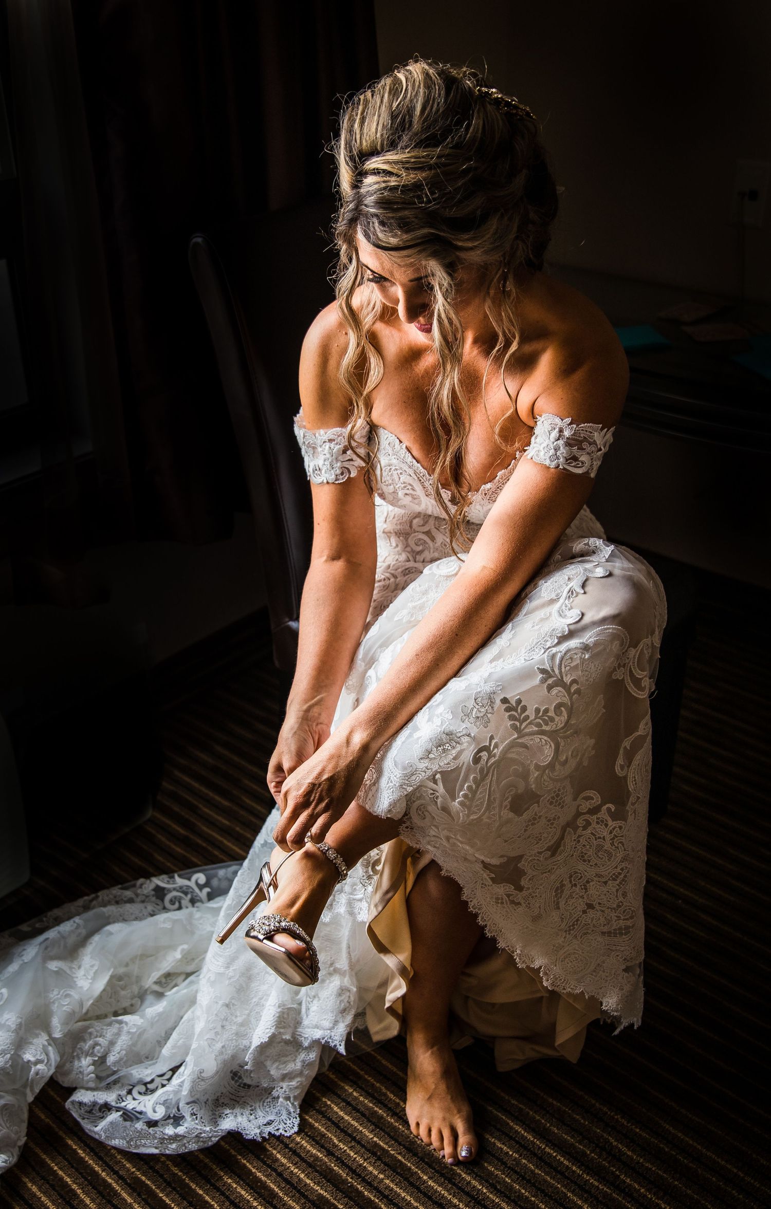 Bride in lace wedding dress adjusts her shoe in dramatic window lighting.