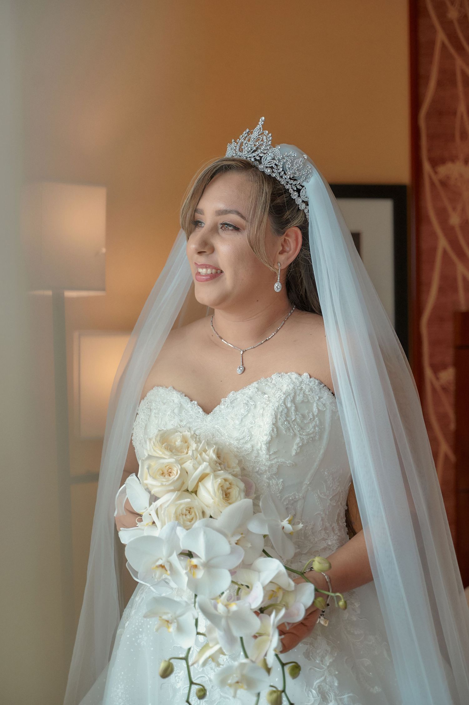 A bride wearing tiara and veil holds white orchid bouquet while smiling.