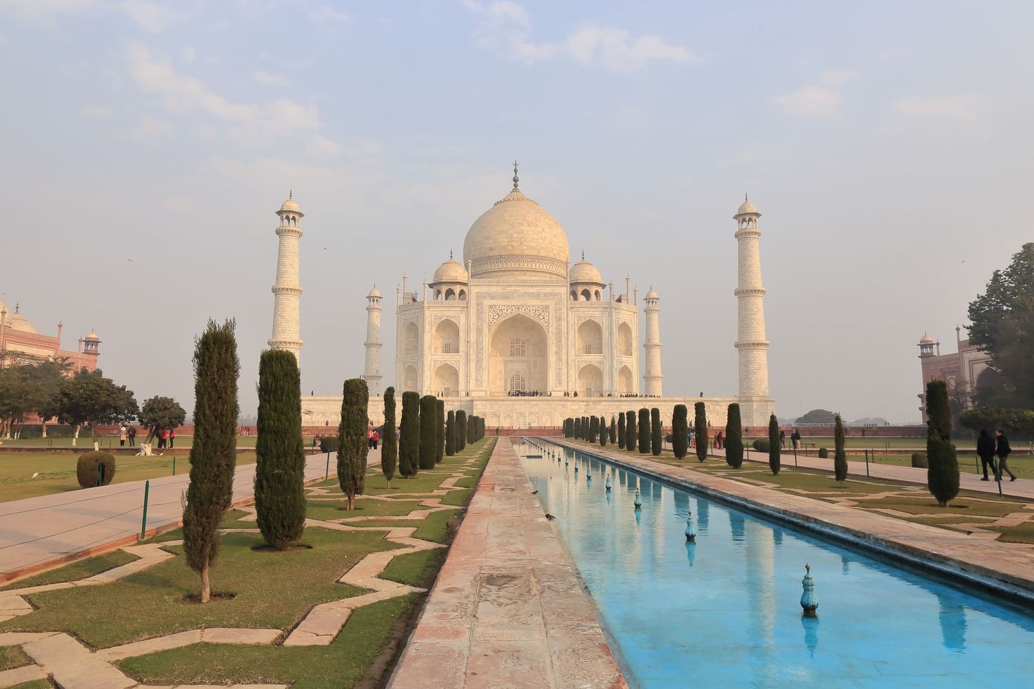 The iconic Taj Mahal with its white marble dome and minarets reflected in a long pool lined with cypress trees.