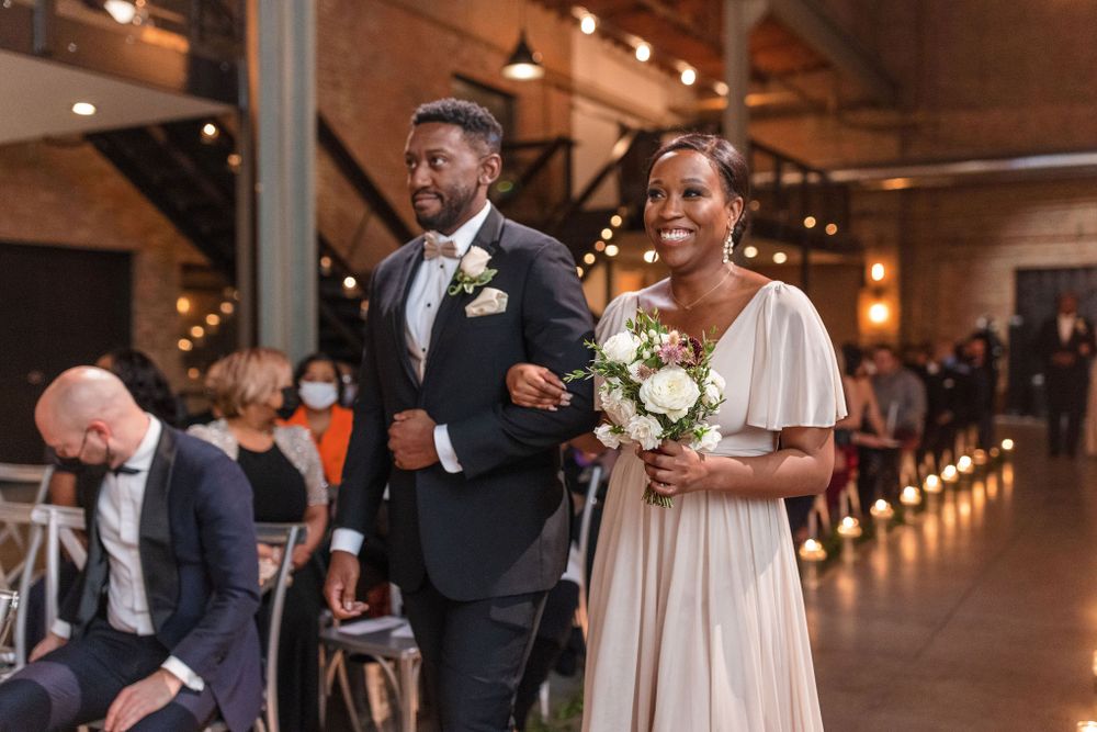 A wedding procession with attendants walking down the aisle at a rustic brick-walled venue with string lights.