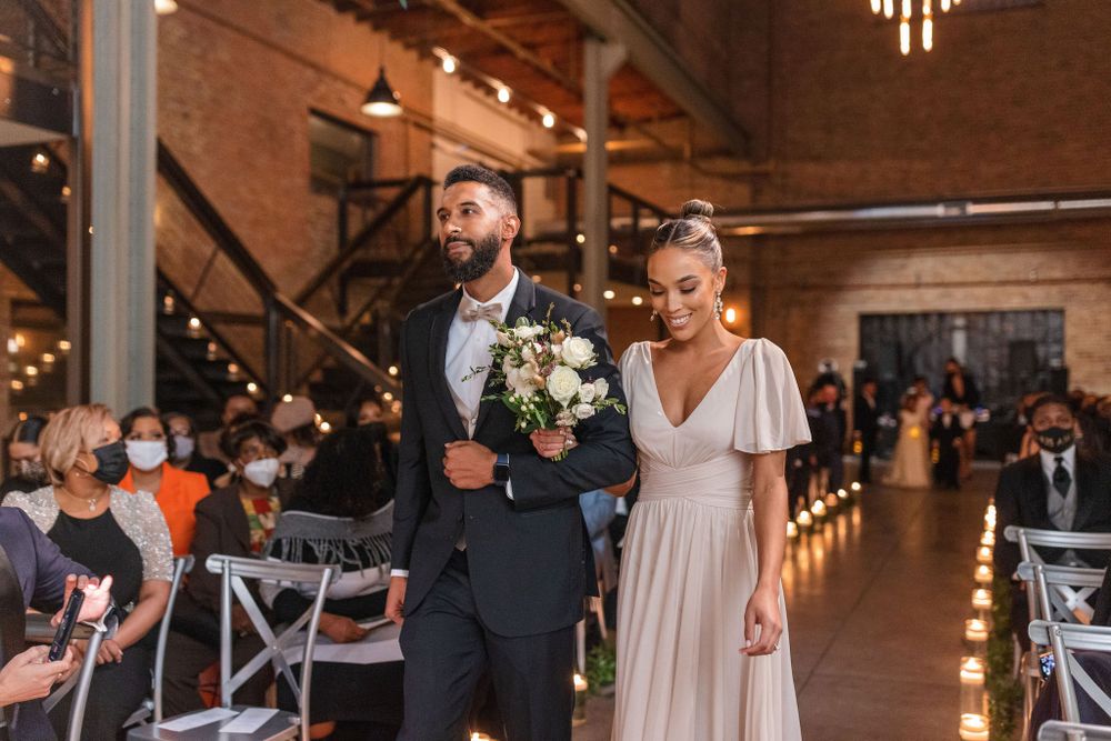 Wedding party members walking down a candle-lit aisle in an industrial chic venue.