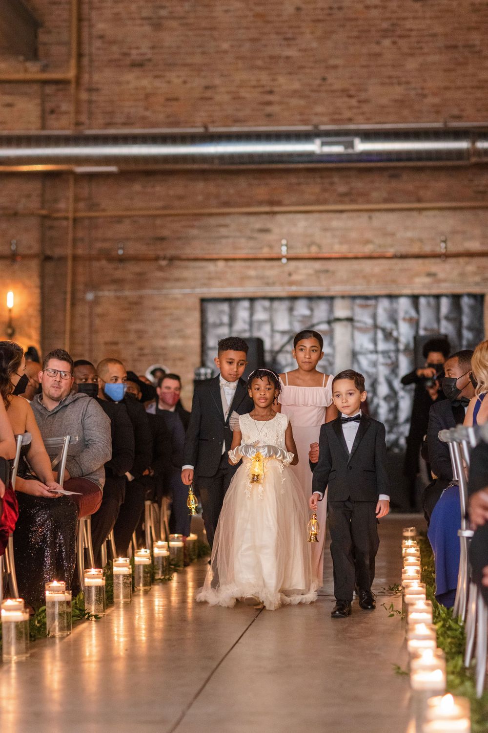 Candlelit aisle walkway with children in formal attire at an indoor wedding ceremony.