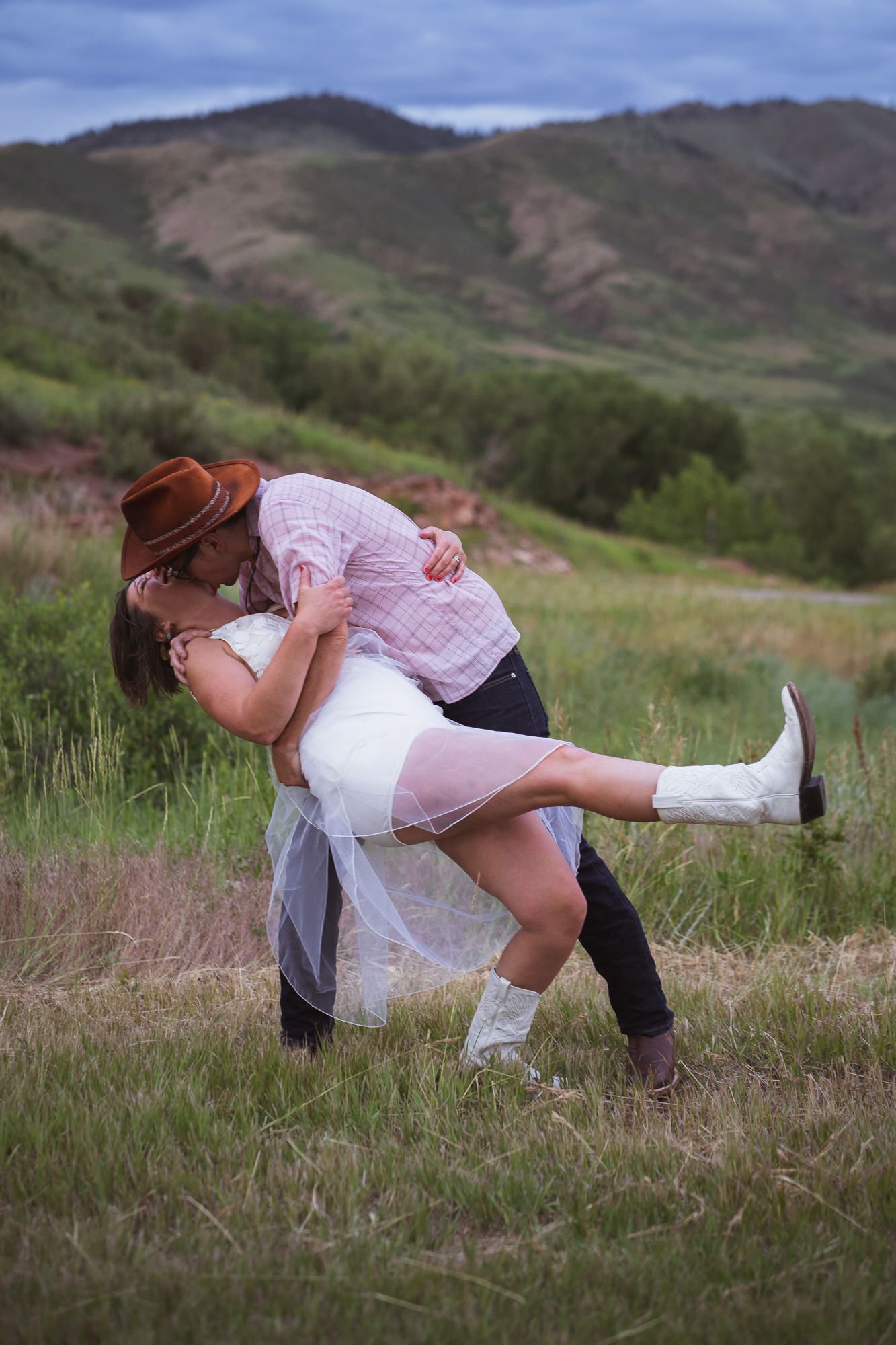 Country western groom dipping and kissing alternative bride wearing white boots