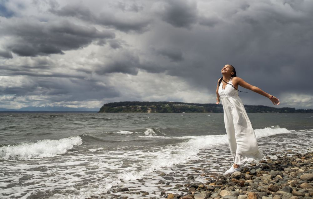 White-dressed figure poses dramatically on rocky beach with stormy skies and waves.