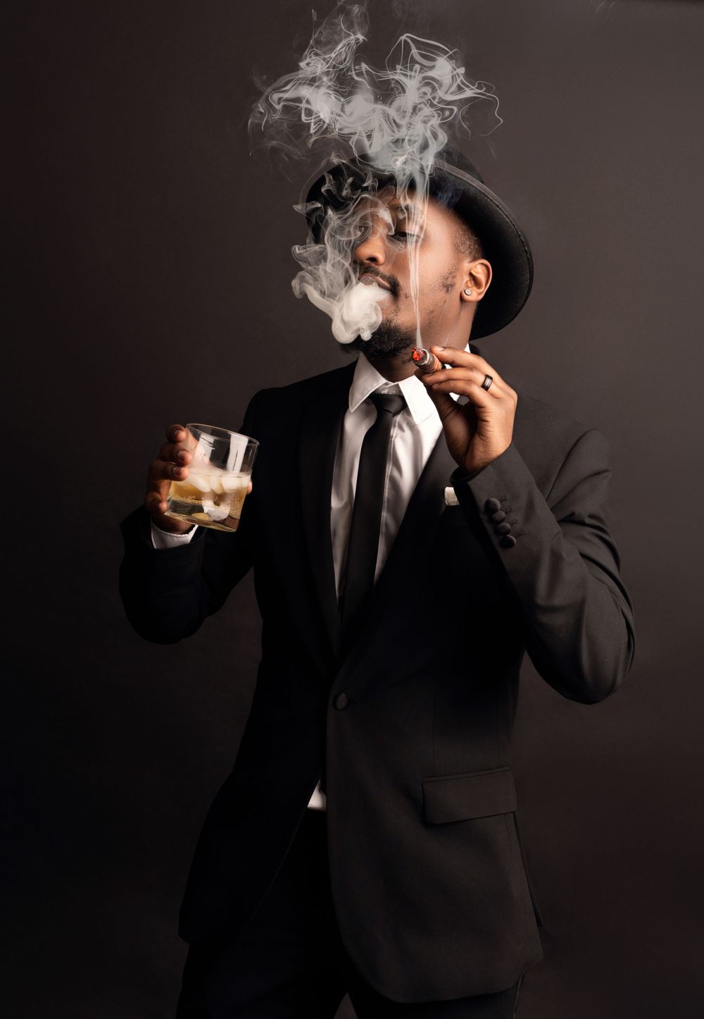 Stylishly dressed figure in black suit exhaling smoke while holding whiskey glass against dark background.