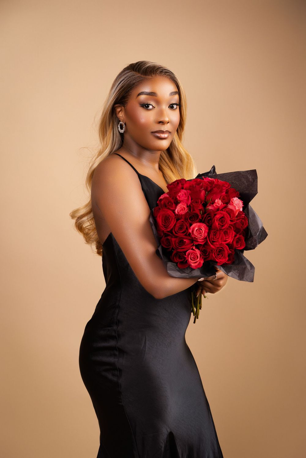Elegant model in black evening dress holding vibrant red rose bouquet against beige backdrop.