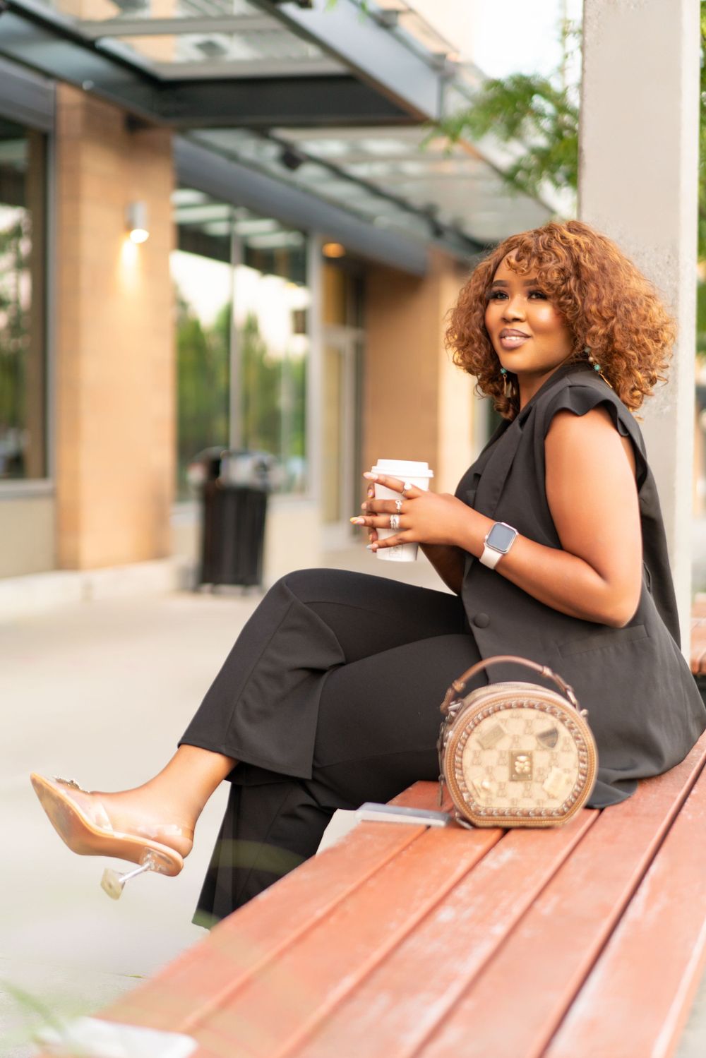 Casual outdoor photo sitting on bench with coffee and designer bag in black outfit.