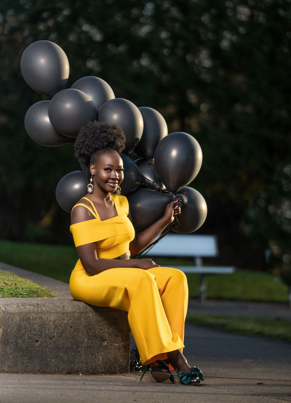 Person in yellow jumpsuit sits outdoors holding black balloons.