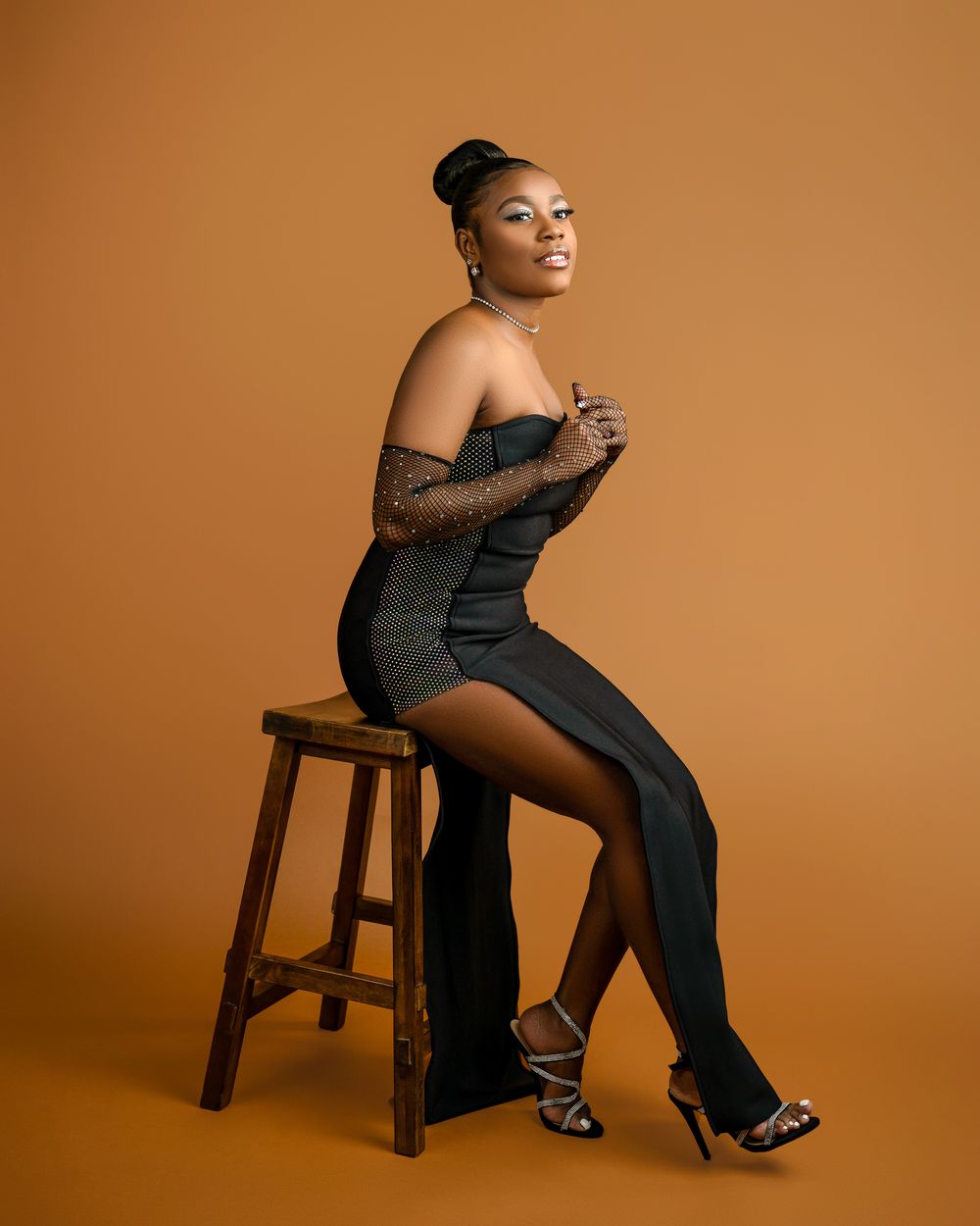 Stylish studio portrait on wooden stool against warm brown background.