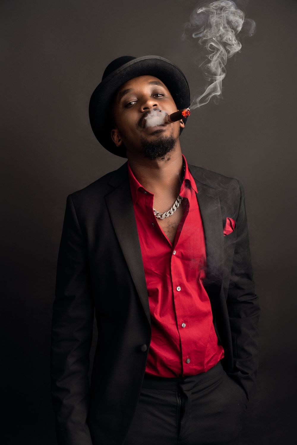 Sophisticated portrait in black suit and red shirt with cigar smoke against dark background.