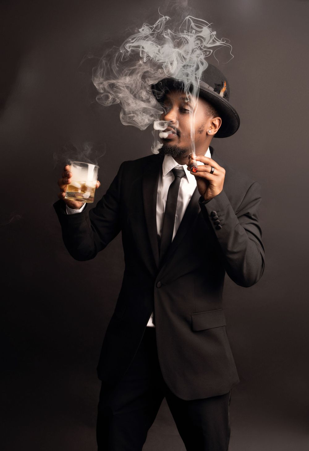 Person in formal attire with fedora blowing smoke rings while holding a drink in dramatic lighting.