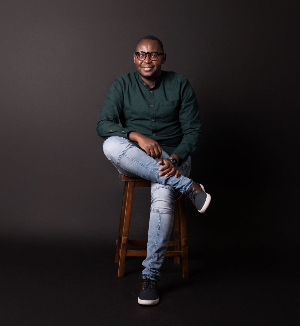 Professional studio portrait of person in dark green button-up shirt and jeans sitting casually on wooden stool.