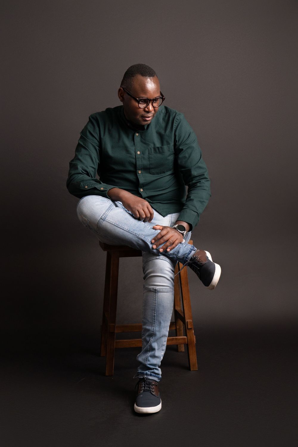 Studio photo showing relaxed pose on wooden stool wearing green shirt and distressed jeans.
