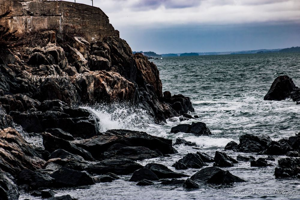 Ocean waves breaking against dark rocky coastline with dramatic cloudy sky.