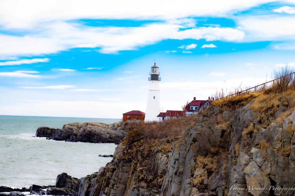 White lighthouse perched on rugged coastal cliffs with bright blue sky and ocean views.