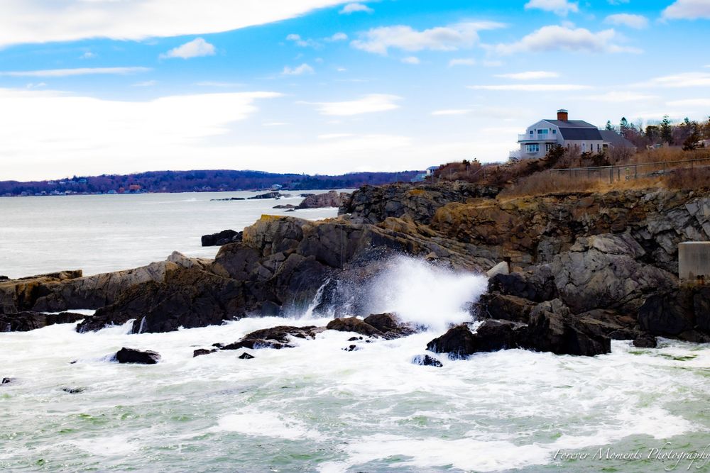 Waves crash against rocky Maine coastline with lighthouse and buildings visible on the cliffs.