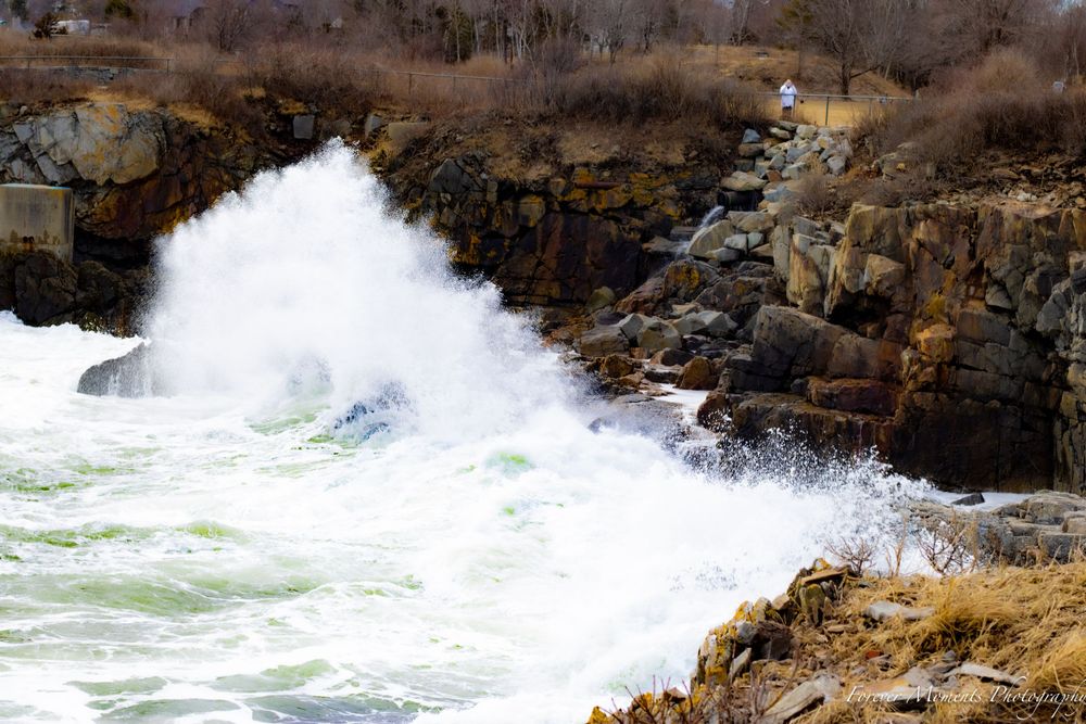 Powerful ocean waves explode against jagged coastal rocks beneath cloudy winter skies.