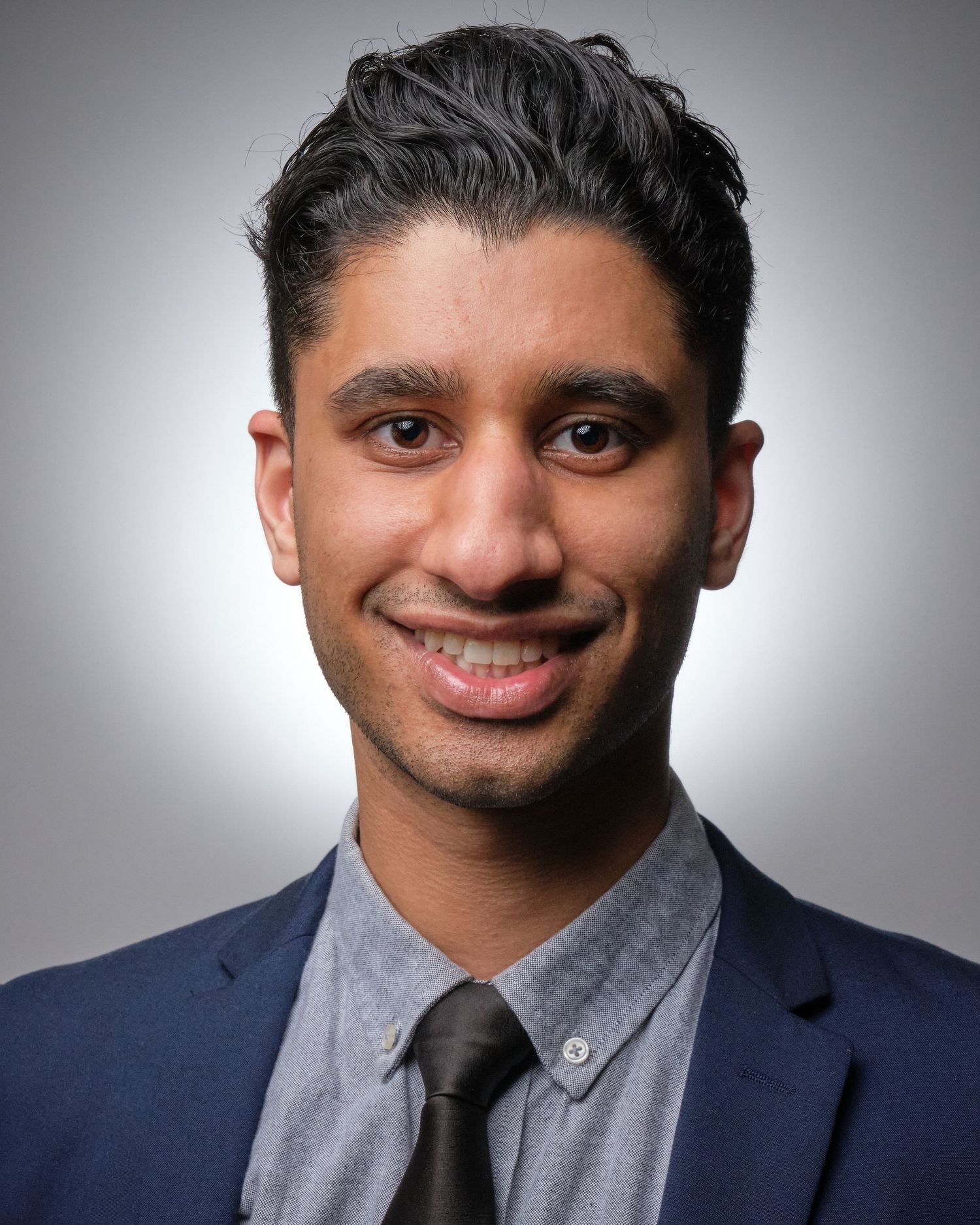 Professional headshot of a person in a navy suit and grey shirt against a gradient background.