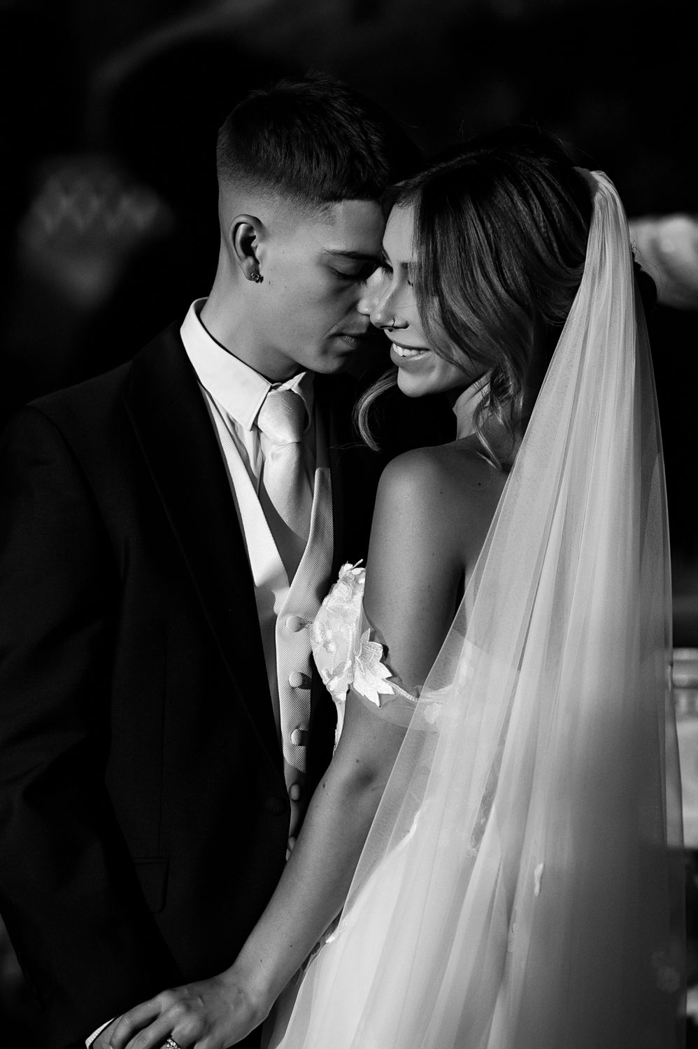 Intimate black and white wedding moment with couple in formal attire and flowing bridal veil.