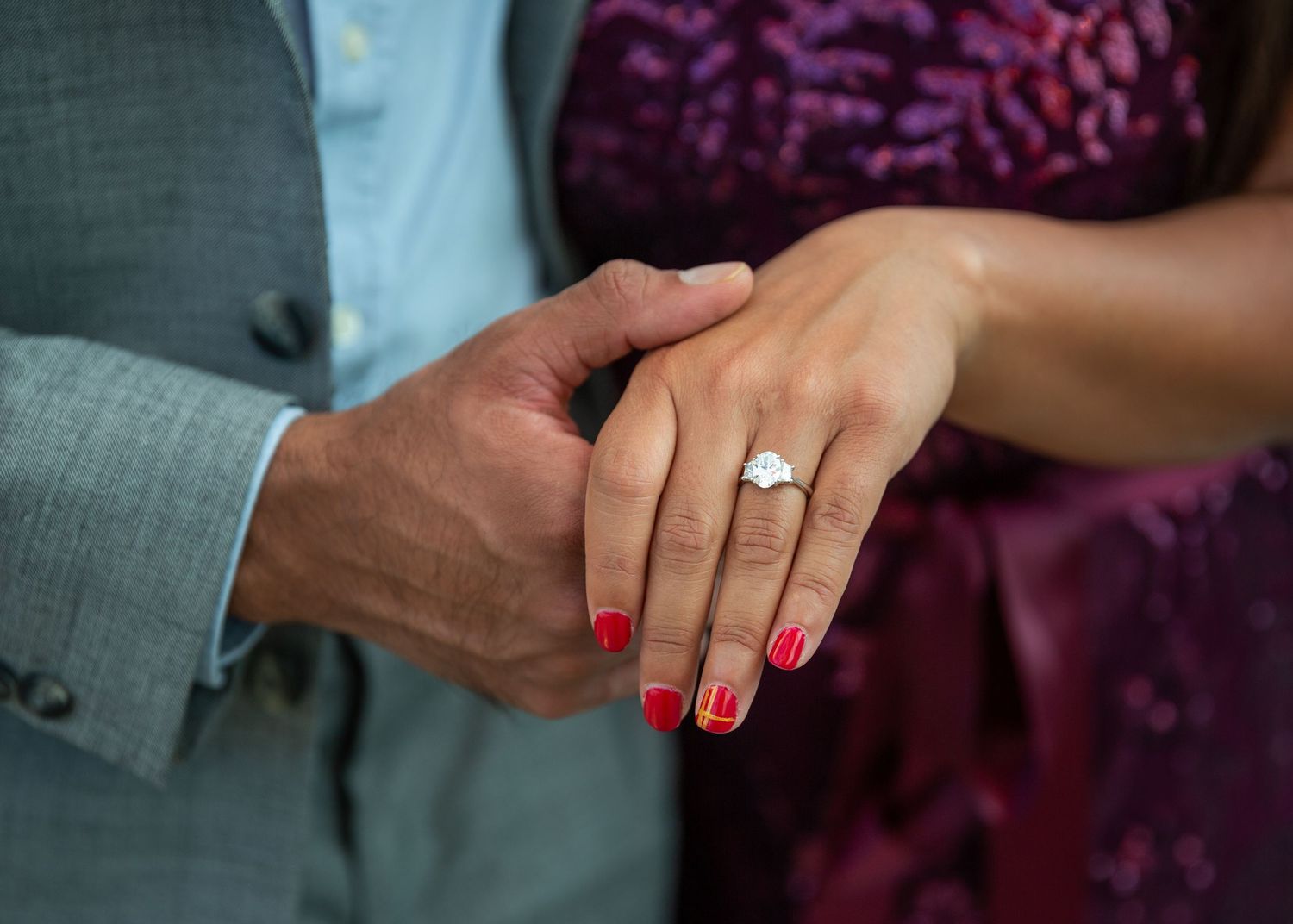 A close-up of hands showing off an engagement ring with red nail polish against formal attire.