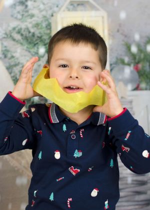 Young boy in Christmas-themed pajamas playfully holds yellow tissue paper to his face.