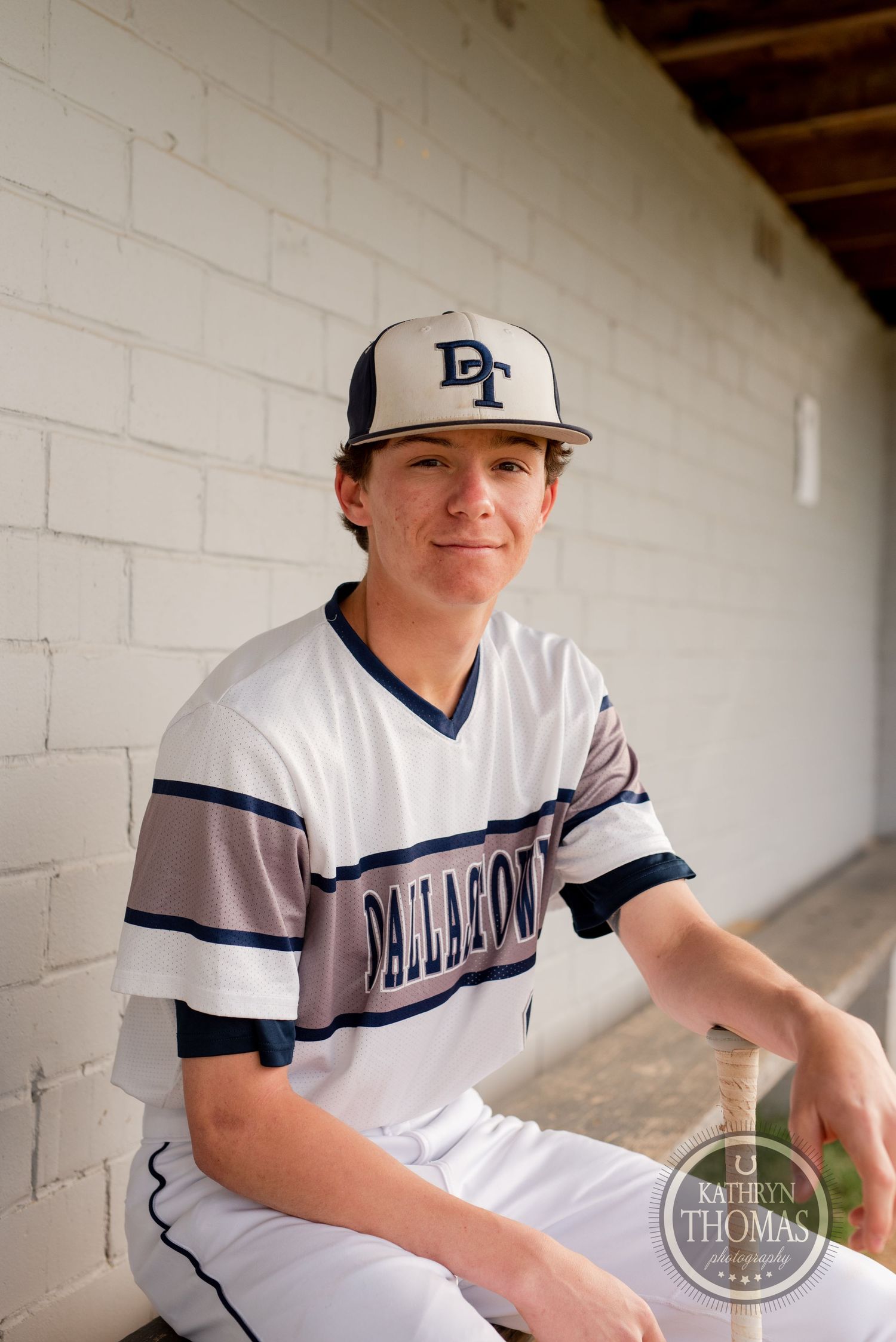 Baseball player in Dellasoft uniform poses in dugout wearing navy and white cap.