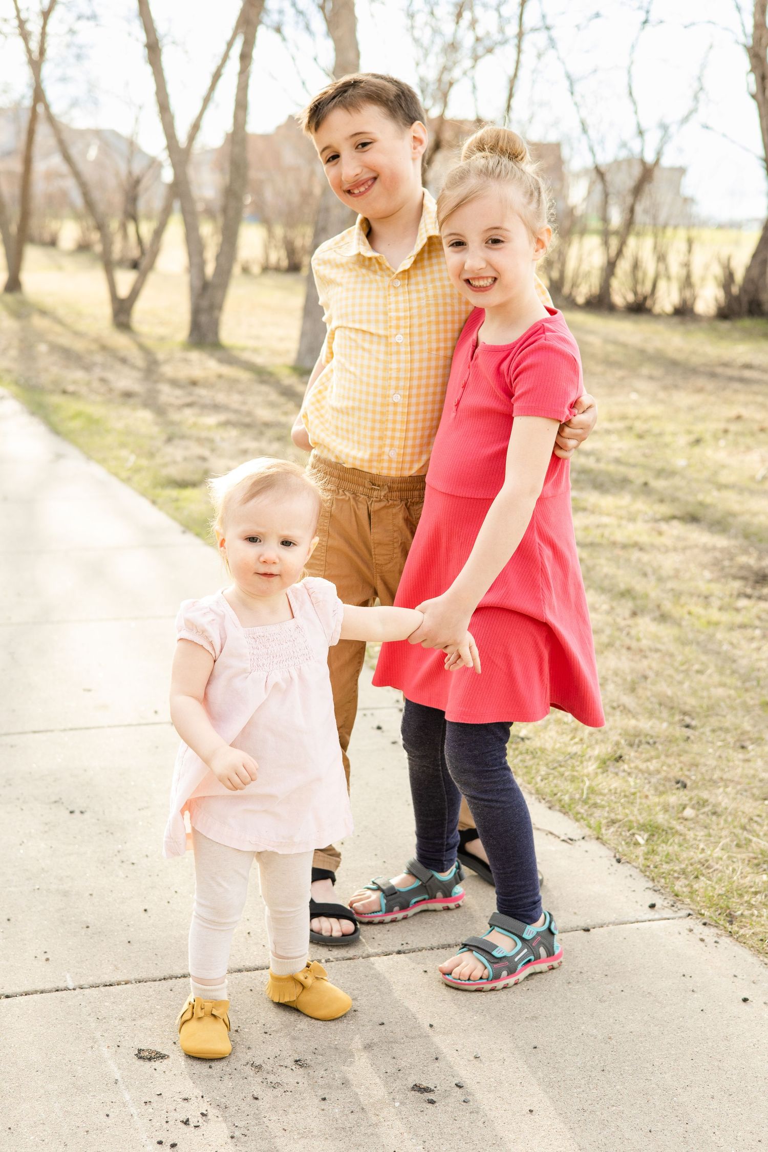 Three siblings stand together on a pathway during a spring day with bare trees in the background.