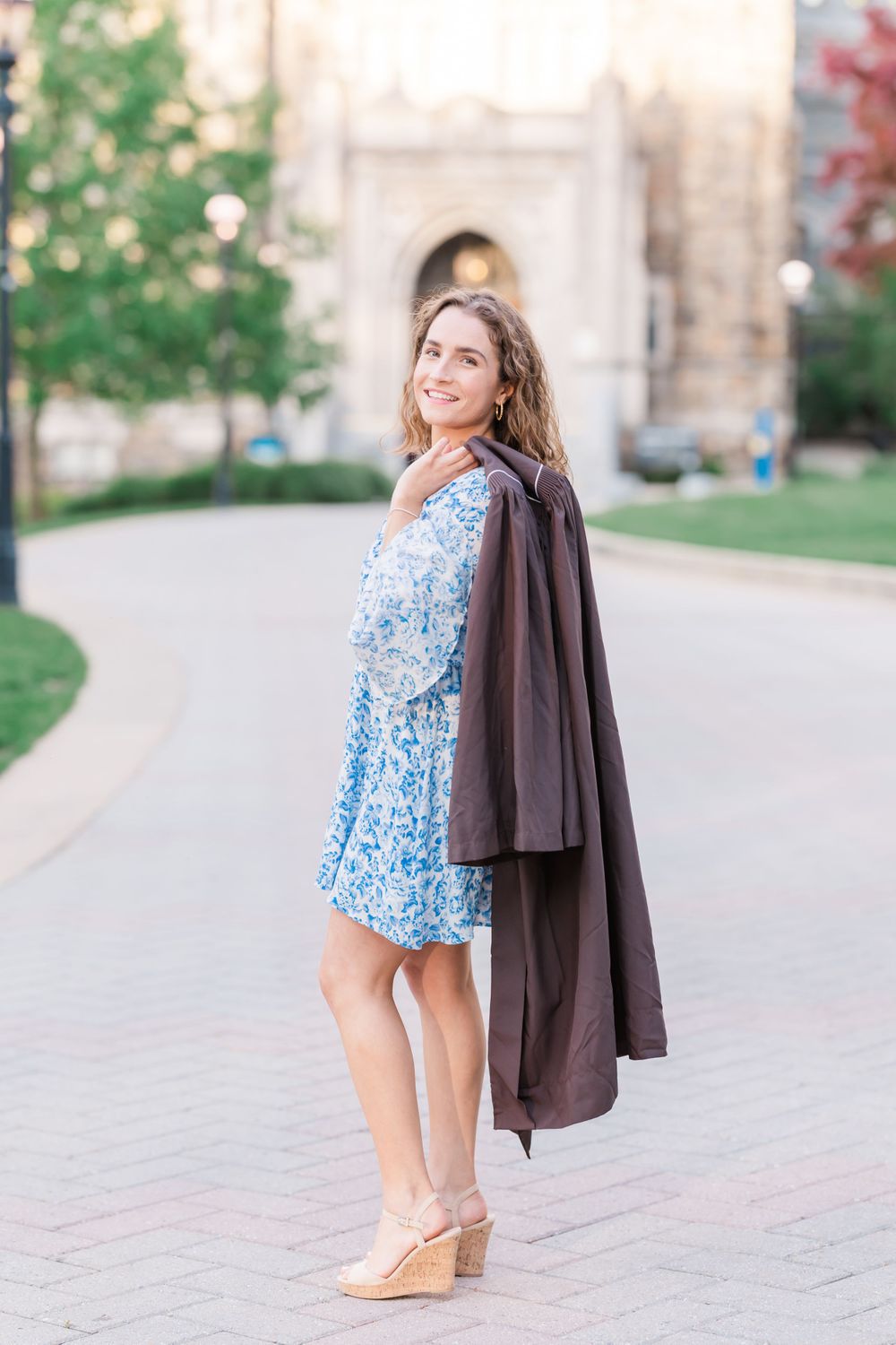 A person in a blue floral dress and brown coat poses on a brick walkway near a stone building.