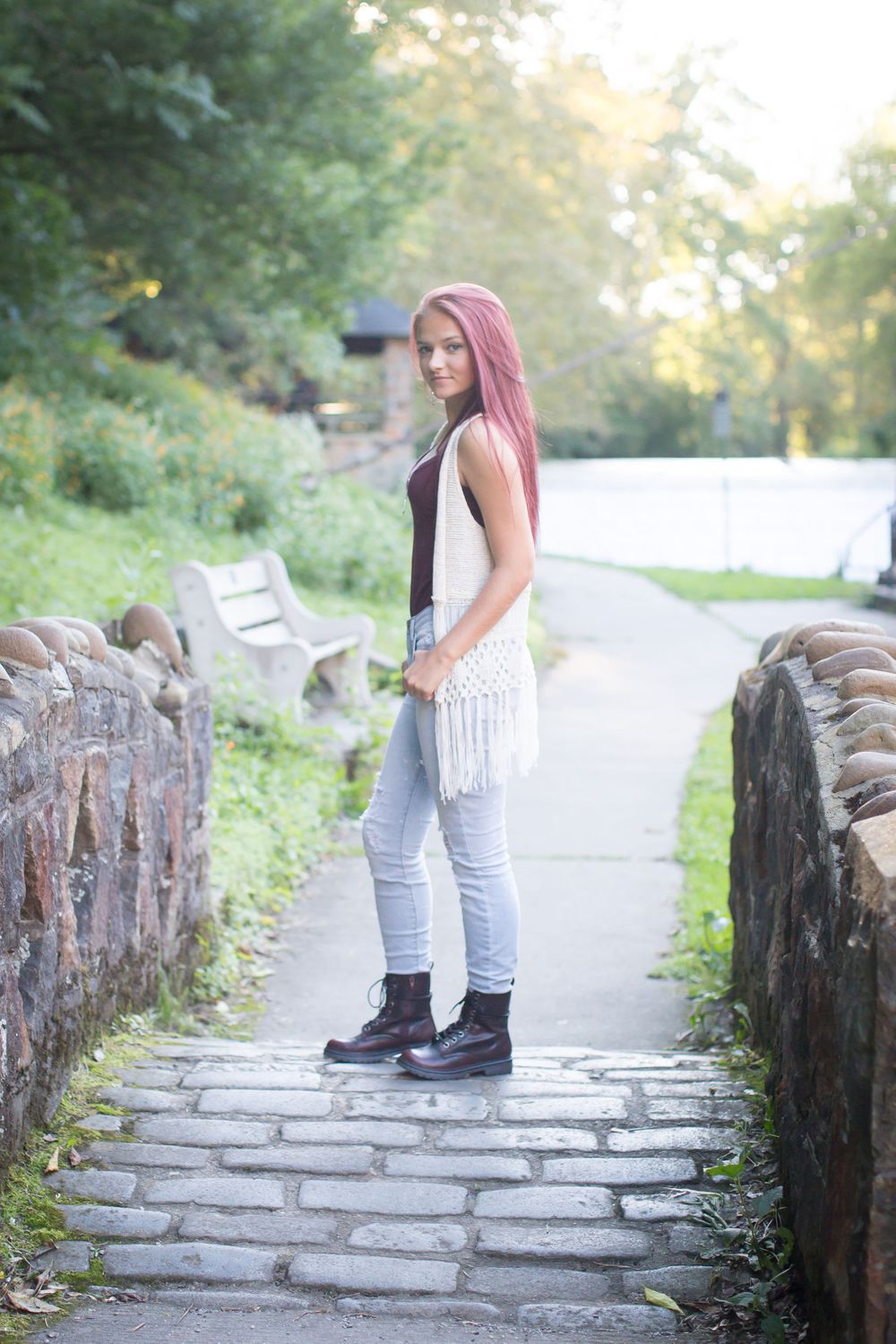 A person wearing a fringe vest and jeans poses on a stone bridge path in a park.