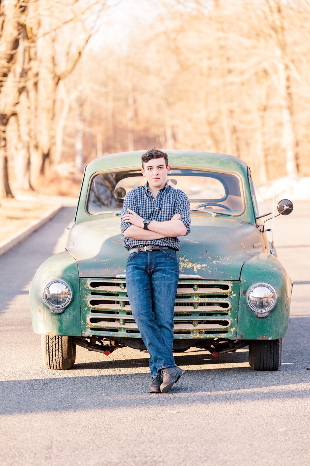 Person in casual attire leaning against vintage green pickup truck on tree-lined road.