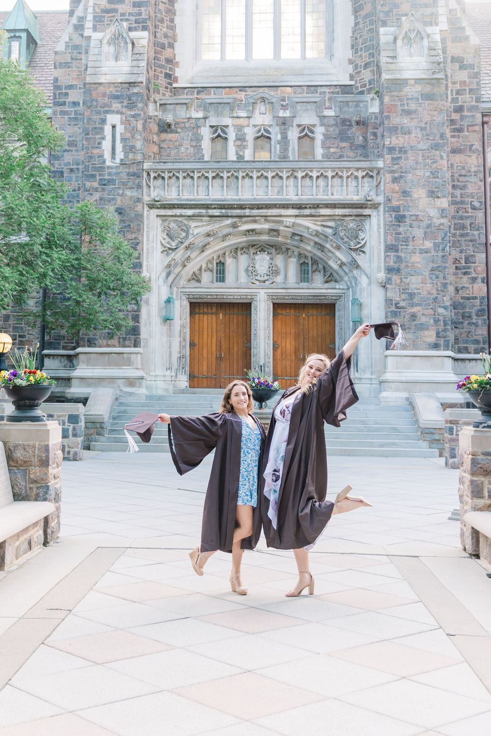 Two people in graduation gowns jump joyfully in front of gothic church doors.