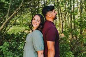 A couple stands back to back smiling in a sun-dappled wooded area on a bright summer day.