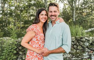 A couple poses together outdoors by a stone wall, with the woman wearing a red floral dress and man in a blue shirt.