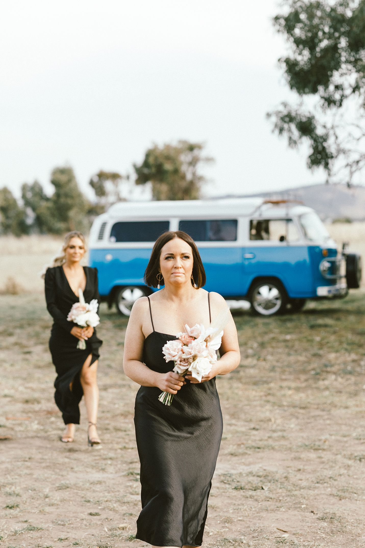 Wedding party members in black dresses walking across outdoor venue with vintage VW bus.