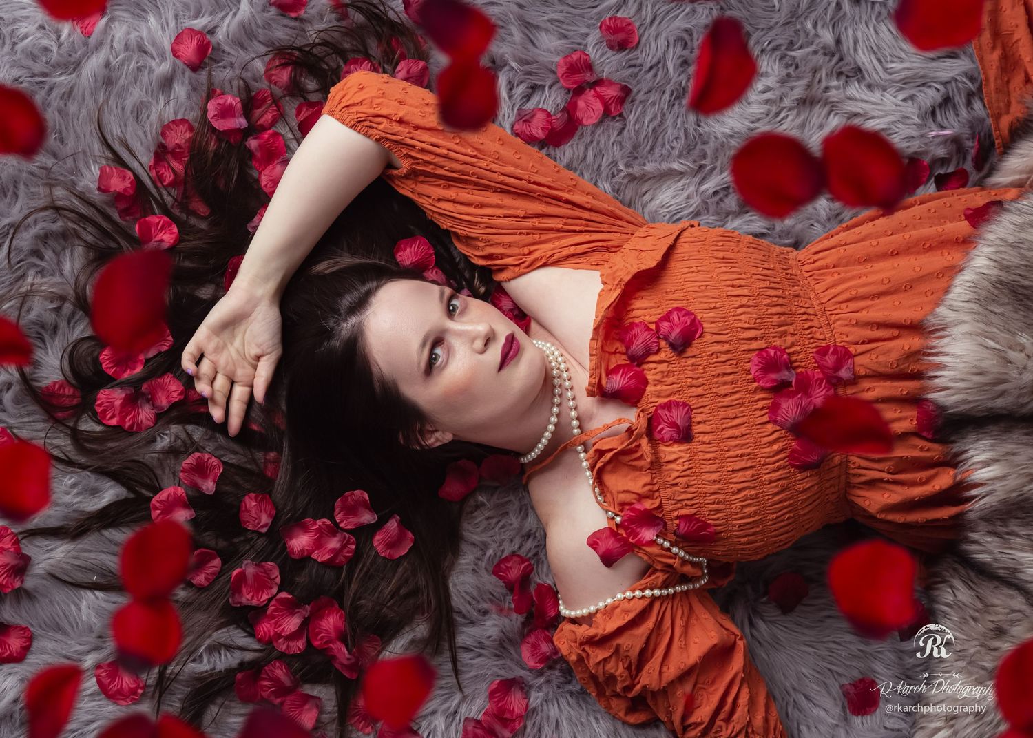Woman in orange dress lying among scattered red rose petals in artistic portrait.