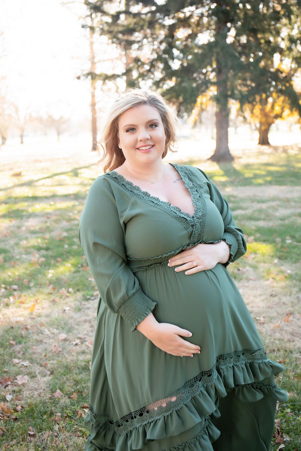 A pregnant woman in a flowing green dress poses in a sunlit park.
