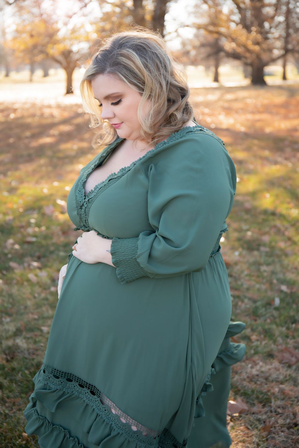 A woman in a green dress cradles her baby bump in an autumn outdoor setting.