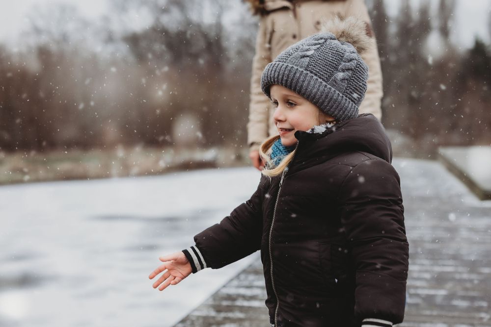 Child in winter clothing experiencing snowfall with arms outstretched.