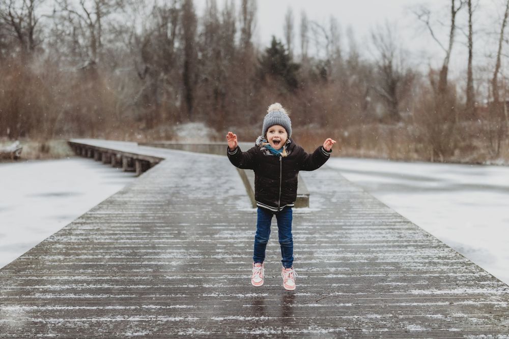 Small figure joyfully poses with arms raised on a wooden boardwalk during winter.