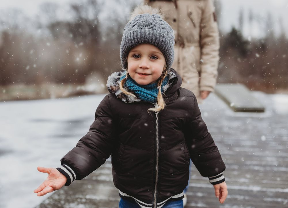 Child in winter coat and knit hat enjoying snowfall outdoors.