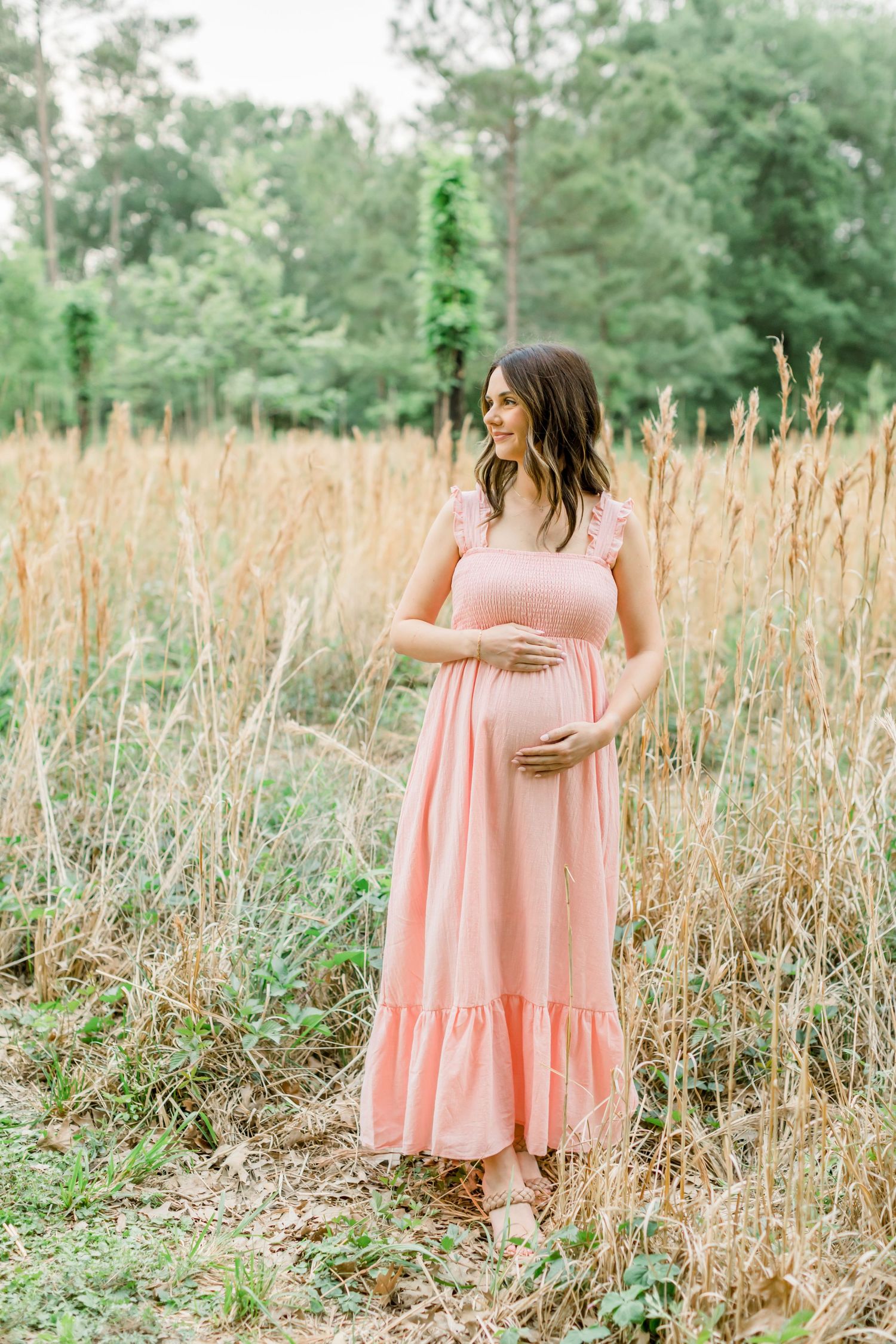 A woman in a pink maxi dress stands alone in a field of tall grass with pine trees in the background.
