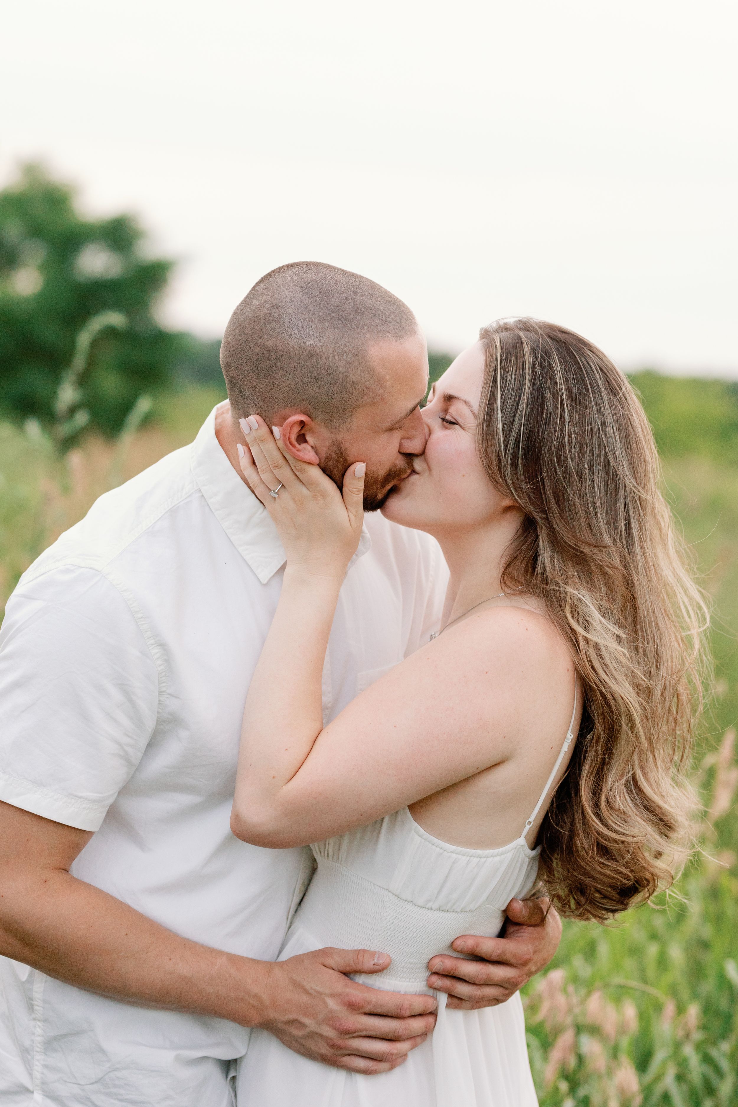Couple embracing and kissing in a sunny field wearing white and casual clothing.