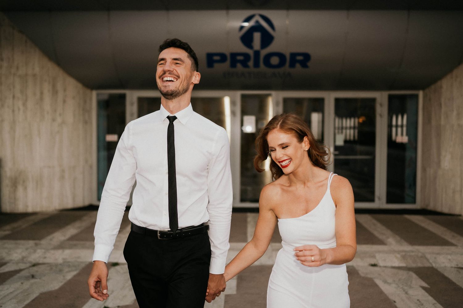 A couple in wedding attire walking hand in hand outside a modern building entrance.