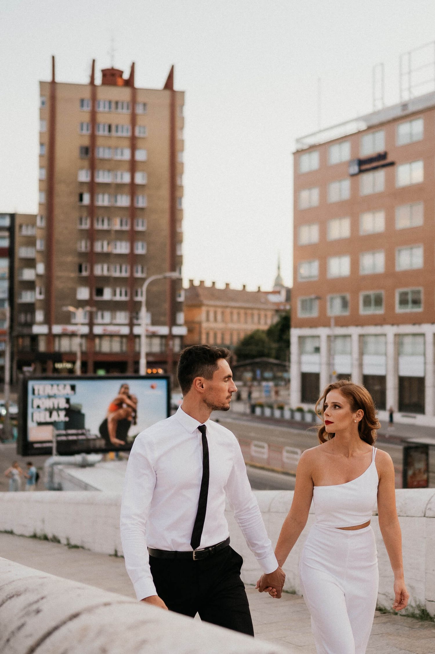 A couple in formal wear walking together against an urban backdrop of tall apartment buildings.