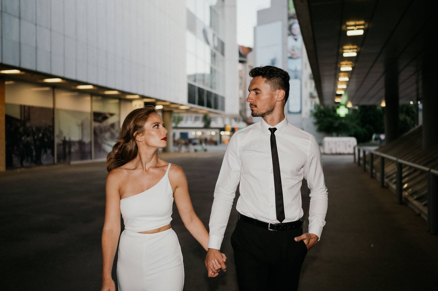 A couple walking hand in hand through an urban corridor wearing coordinated formal attire.